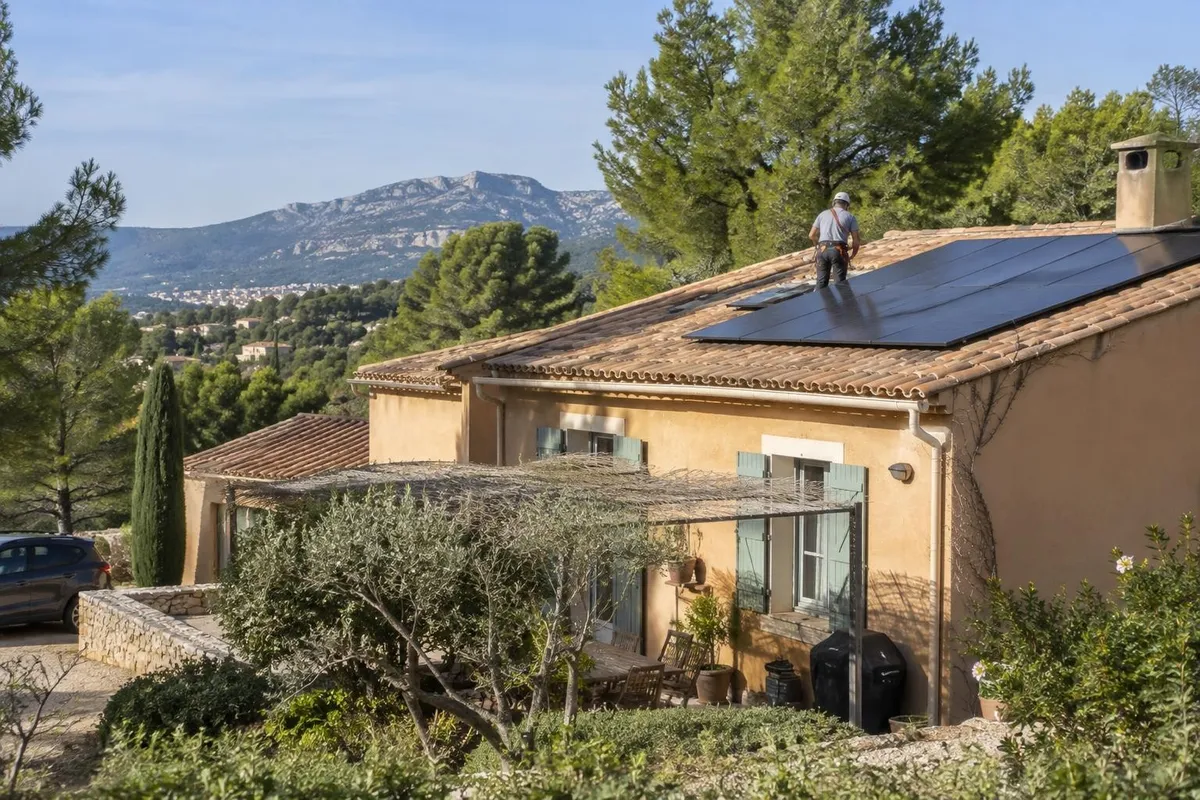 Maison provençale aux tuiles en terre cuite équipée de panneaux solaires, avec un électricien vu de loin sur la toiture.